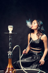 Fototapeta na wymiar Young beautiful female in black dress smoking and exhales hookah
