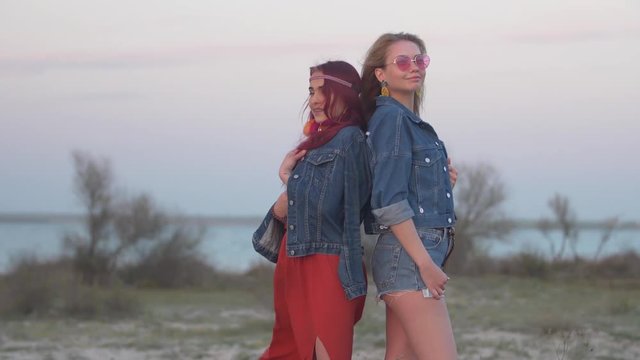two young women dressed in denim jackets stand back to back in the gentle rays of the sunset with hair flying in the wind against the lake