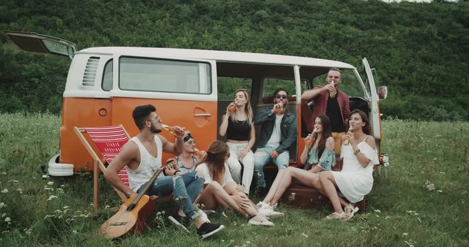 Big group of friends at the picnic party drinking beer and cheers, sitting behind of a retro van