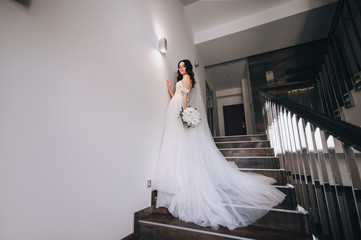 A beautiful and sweet bride in a white wedding and long dress is standing on an old staircase....