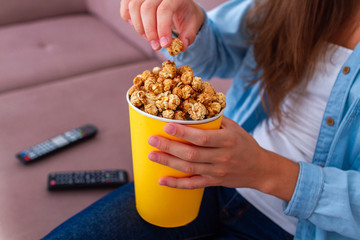 Woman sitting on couch and eating crunchy caramel popcorn during watching tv at home
