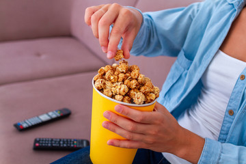 Obraz premium Woman sitting on couch and eating crunchy caramel popcorn for snack during watching tv at home