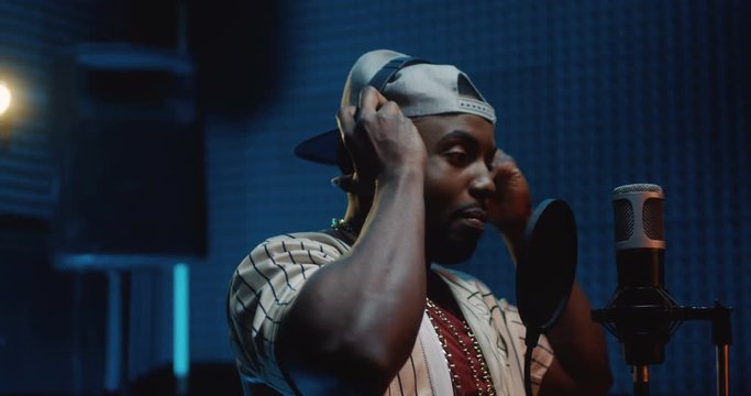 Handsome and hapy young African American guy in a hat singer taking on headphones and recording a song in the dark sound studio. Close up.