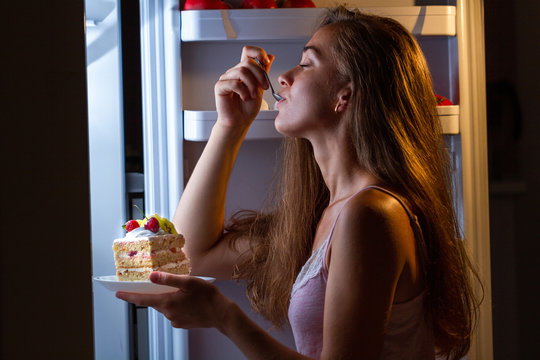 Hungry Woman In Pajamas Enjoys Sweet Cake At Night Near Refrigerator. Stop Diet And Gain Extra Pounds Due To Carbs Food And Unhealthy Eating