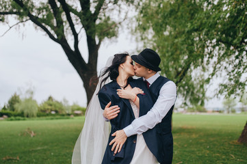 Naklejka premium Beautiful newlyweds are hugging against the background of green grass and willows in the park and garden. Wedding portrait of a stylish groom in a hat and a beautiful bride with curly hair in a dress.