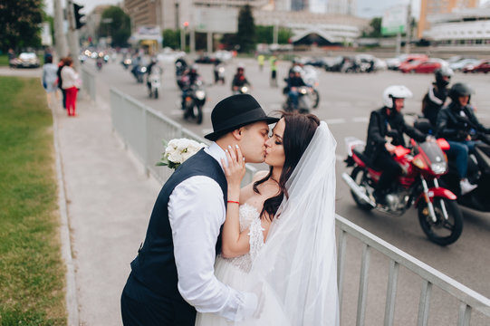 Stylish groom ganster in a suit and a hat embraces and kisses a young and beautiful bride against the background of motorcyclists and motorcycles. Wedding portrait.