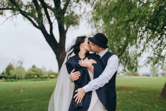 Beautiful newlyweds are hugging against the background of green grass and willows in the park and garden. Wedding portrait of a stylish groom in a hat and a beautiful bride with curly hair in a dress.
