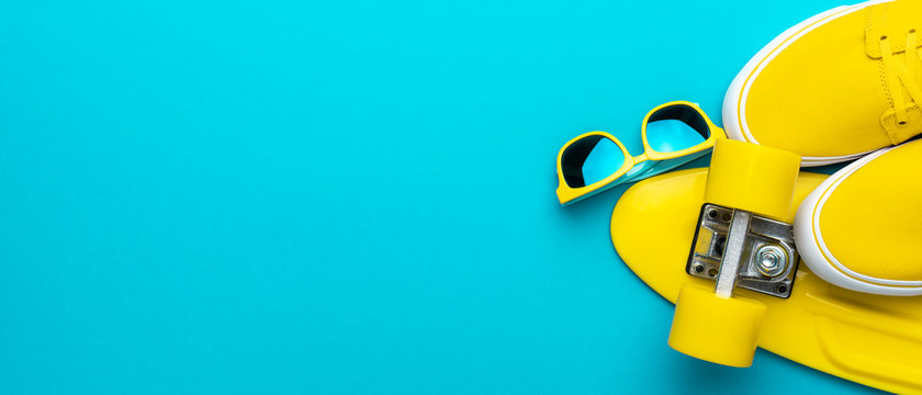 Top view panoramic image of yellow modern teenage accessories. Flat lay photo of yellow sunglasses, sneakers, plastic mini cruiser skateboard over blue turquoise background with copy space.
