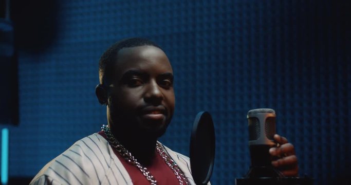 Young African American attractive man singing a song in the microphone at the sound studio, then taking on a cap and smiling t the camera. Close up. Portrait.