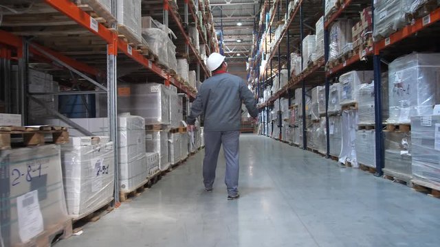 An employee at a logistics warehouse checks products. A man walks along huge shelves with boxes. 4K Slow Mo