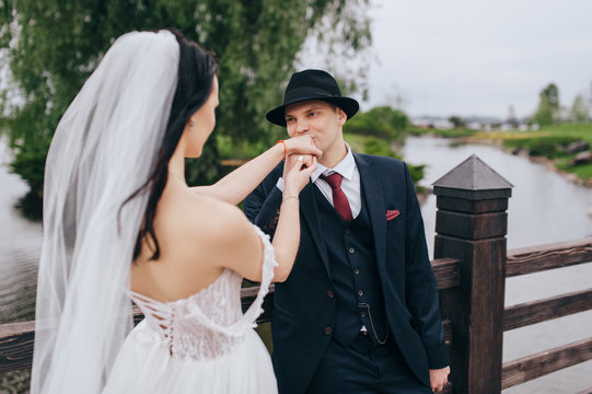 Beautiful newlyweds hugging and kissing on an old bridge, against the backdrop of nature and the lake. Wedding portrait of a stylish groom in a hat who kisses the hand of a brunette bride.