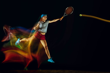 Fototapeta premium One caucasian woman playing tennis isolated on black background in mixed and stobe light. Fit young female player in motion or action during sport game. Concept of movement, sport, healthy lifestyle.