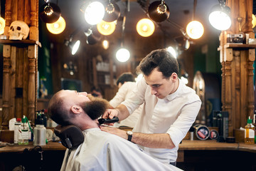 Pleasant hardworking barber holding electric trimmer for shaving and cutting beard. Fashionable beard styling. Barbershop interior with elements of eclecticism and creative lighting. Side view.