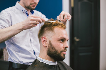 Barber Making Haircut Bearded Man In Barbershop. Professional stylist cutting client hair in salon. Barber using scissors and comb. Skillful hairdresser cutting male hair. Hair Care Service Concept