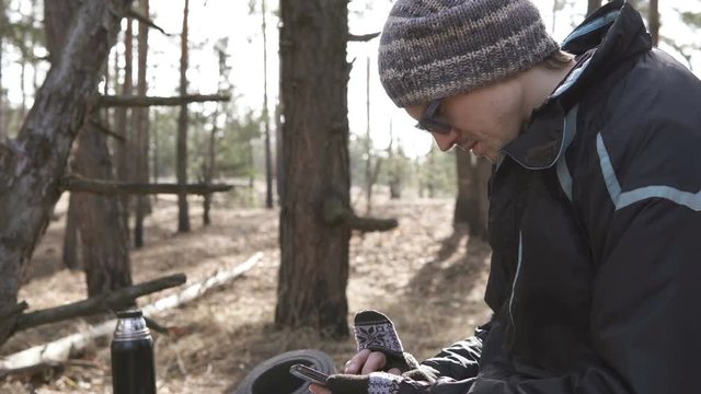 A tourist in the forest looks at a map on a smartphone and drinks tea from a thermos