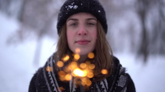 Portrait of cute smiling young woman in warm jacket with burning sparkler in winter park. Focus changing from face to sparks. Winter leisure. Slow motion