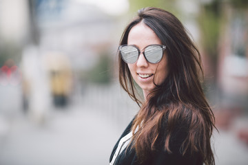 Portrait of a young beautiful caucasian woman on the background of the street. Model girl with long hair and sunglasses with reflection posing in black clothes in an urban environment