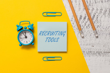 Text sign showing Recruiting Tools. Business photo text getting new talents to your company through internet or ads Notepad clips colored paper sheet markers alarm clock wooden background