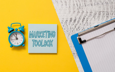 Conceptual hand writing showing Marketing Toolbox. Concept meaning Means in promoting a product or services Automation Notepad colored paper sheet alarm clock wooden background
