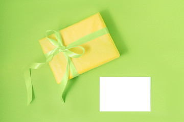 Yellow gift box with green silk ribbon and empty isolated label on pastel green background. Holiday gift concept. Flat lay. Copy space.