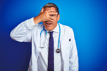 Handsome middle age doctor man wearing stethoscope over isolated blue background peeking in shock covering face and eyes with hand, looking through fingers with embarrassed expression.