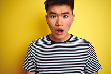 Young asian chinese man wearing striped t-shirt standing over isolated yellow background scared in shock with a surprise face, afraid and excited with fear expression