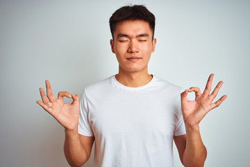 Young asian chinese man wearing t-shirt standing over isolated white background relaxed and smiling with eyes closed doing meditation gesture with fingers. Yoga concept. - Powered by Adobe
