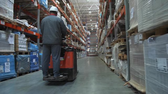 A huge warehouse with goods. The worker in uniform and helmet moves on the square between the rows. 4K Slow Mo
