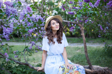 Fototapeta premium Happy Woman Life Style, portrait of a beautiful girl in a straw hat sitting near a blossoming lilac in the rays of the setting sun, picnic. Summer picnic and weekend break concept. Vintage fashion
