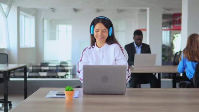 Young Girl Listening To Music With Blue Headphones In Office.Sitting At Table. Work Break. Colleagues On Background.