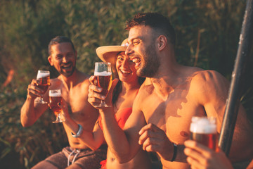 Happy group of young people drinking beer on a dock by the river during the summer sunny day