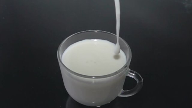 Butter milk (kefir, cultured yoghurt) flows into a transparent cup that stands on a black surface, dairy products, sour milk products, healthy diet. Super slow motion 1000 fps