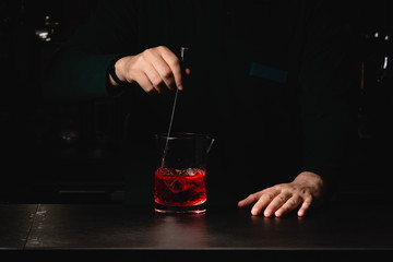 Closeup shot of bartender hand stirring a negroni cocktail. Glass of drink on counter with orange peal