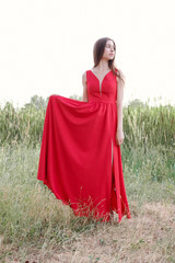 Brown-haired barefoot girl with long hair in red dress on river coast.