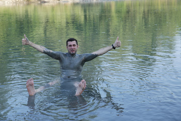 A young handsome man without a shirt, his body smeared with curative mud, sits and relaxes in the water of a mountain river. Healthy lifestyle, natural procedures. The concept of unity with nature.