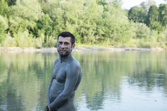 Close-up. A young handsome man without a shirt, his body daubed smeared with therapeutic curative mud, stands in the water of a mountain river. Healthy lifestyle, natural procedures. 