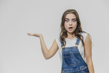 Young student girl standing on a white background. Dressed in white t-shirt and denim overalls. During this time, put one hand to one side. From the surprise of the doorway and looking into the camera