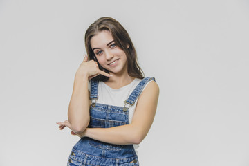 Portrait of a young, sweet, smiling girl in a white t-shirt and denim overalls. Shows gesturing smart phone on one side, standing above a gray background.