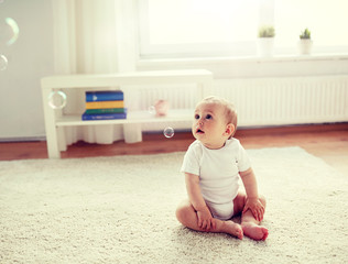 childhood, babyhood and people concept - happy little baby boy or girl sitting on floor with soap bubbles around at home