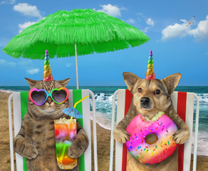 The dog unicorn with a donut and the cat in sunglasses with a glass of colored cocktail are sitting under the green straw umbrella on a beach chairs on the sea shore.