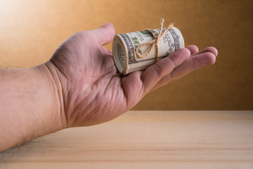 Fototapeta premium A man Hand and a roll of American currency (USD, American dollars) with 100 dollars bank notes on the outside as a symbol of plenty of money on the wooden background. 