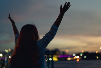 Silhouette behind a young girl raising her hands in the air during golden hour - Woman traveller enjoying watching a beautiful sunset with city bokeh lights - Travel, freedom, lifestyle concept