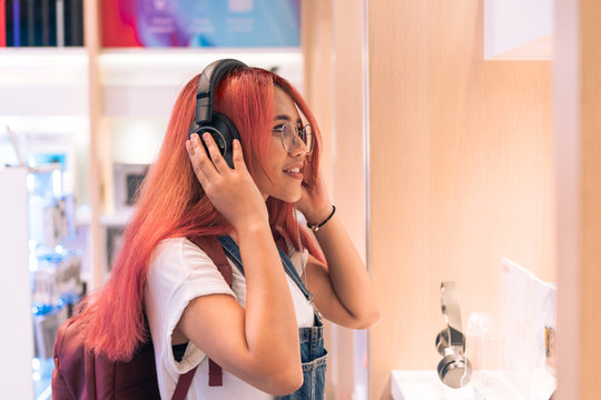 Asian social influencer woman trying on headphones inside retail store - Happy millennial diverse girl shopping and testing lifestyle music tech products - Technology, electronic and purchase concept.