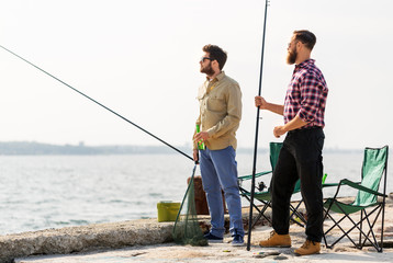 leisure and people concept - male friends with fishing rods and beer on pier at sea