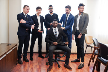 Six multiracial business mans standing at office and roll man on chair. Diverse group of male employees in formal wear having fun.