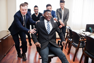 Six multiracial business mans standing at office and roll man on chair. Diverse group of male employees in formal wear having fun.