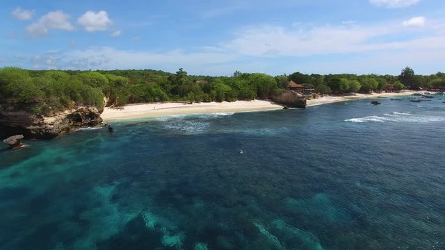 Flying along secret deserted beach between cliffs and lush foliage on bright sunny day. Foamy waves of turquoise Indian Ocean wash the white sand. Aerial of Mushroom Beach, Nusa Lembongan, Bali.