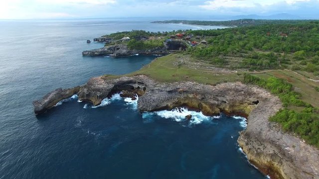 Flying back of rocky ledges with green vegetation, village houses, hiking paths on top. Grottoes and lagoons washed by waters of turquoise ocean. Foamy waves break on stones. Aerial of Nusa Ceningan.