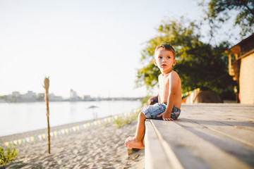 Little Caucasian boy child sitting on wooden pier sandy beach, summer time, sea vacation near...