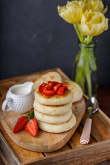 mini stack of cheesecakes on wooden tray, with slices of strawberries, sauce, vase with tulips on table
